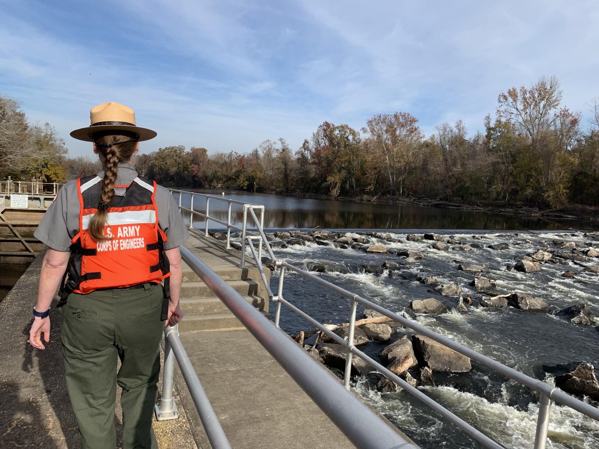 woman walks away from camera with long braided hair in green pants, orange lifejacket, and tan hat. walking atop a concrete ledge overlooking rapids in the water