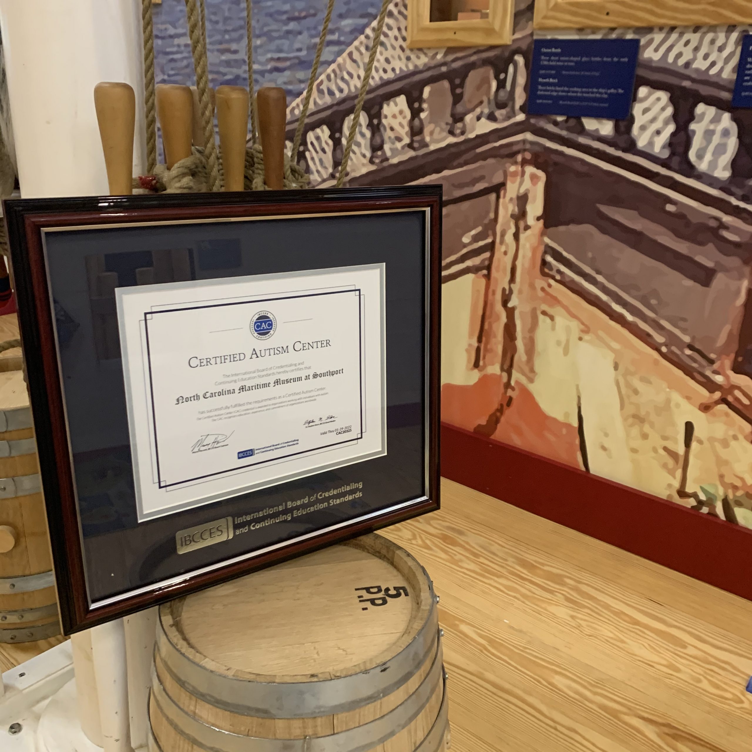 dark frame and matting surround a white with blue accent certificate leaned on top of a brown barrel and leaned against a white pillar in wooden floors and boat covered wall
