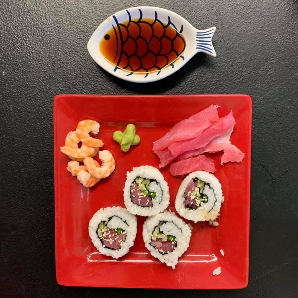 red square plate with four cooked shrimp in top left corner with green wasabi and pink ginger to its right and four rolled sushi rings across bottom
