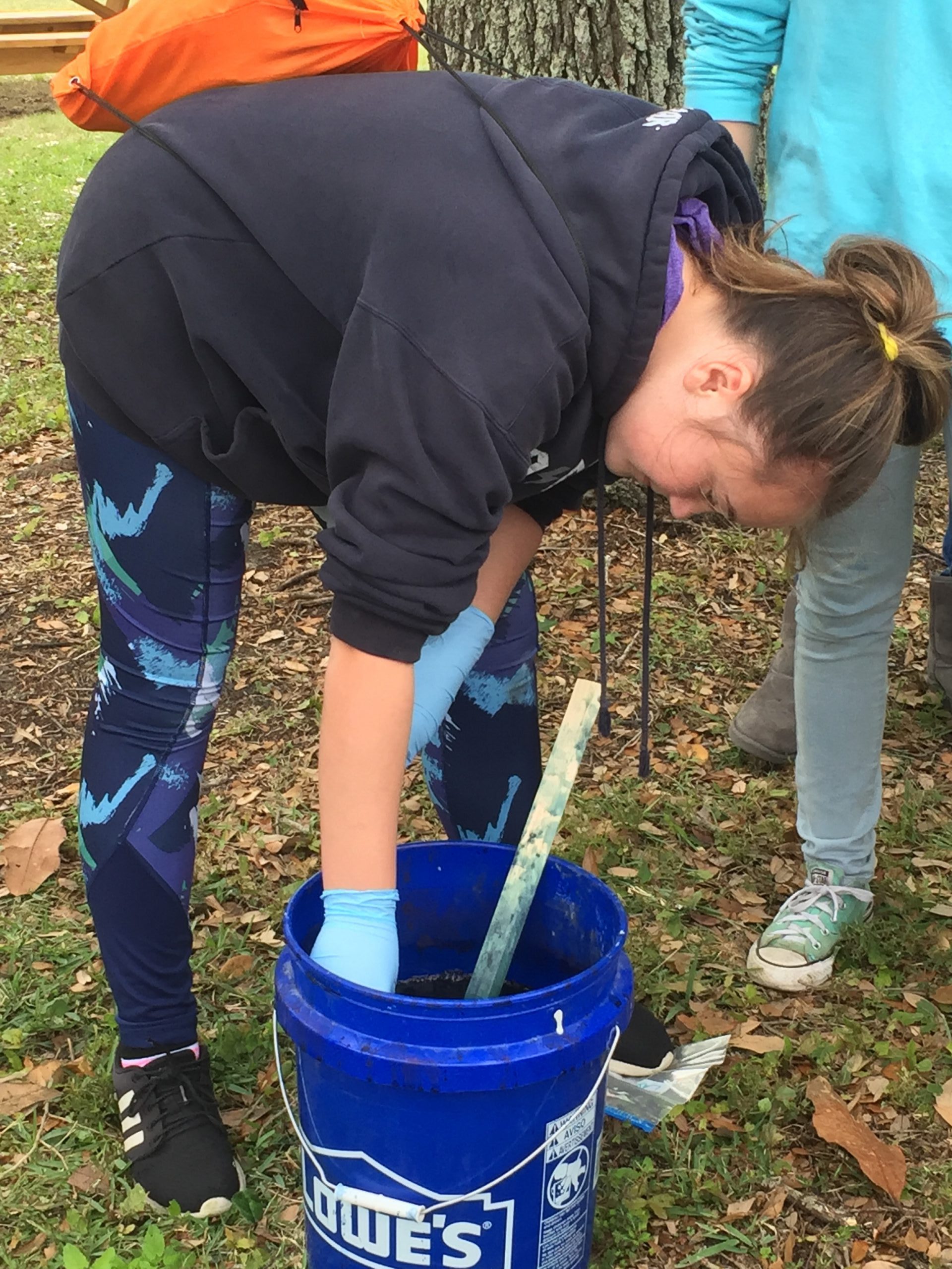 caucasian teenager with brown hair in bun wearing blue leggings and a blue hoodie bends over blue bucket with wooden stirer wearing blue gloves while standing on grass
