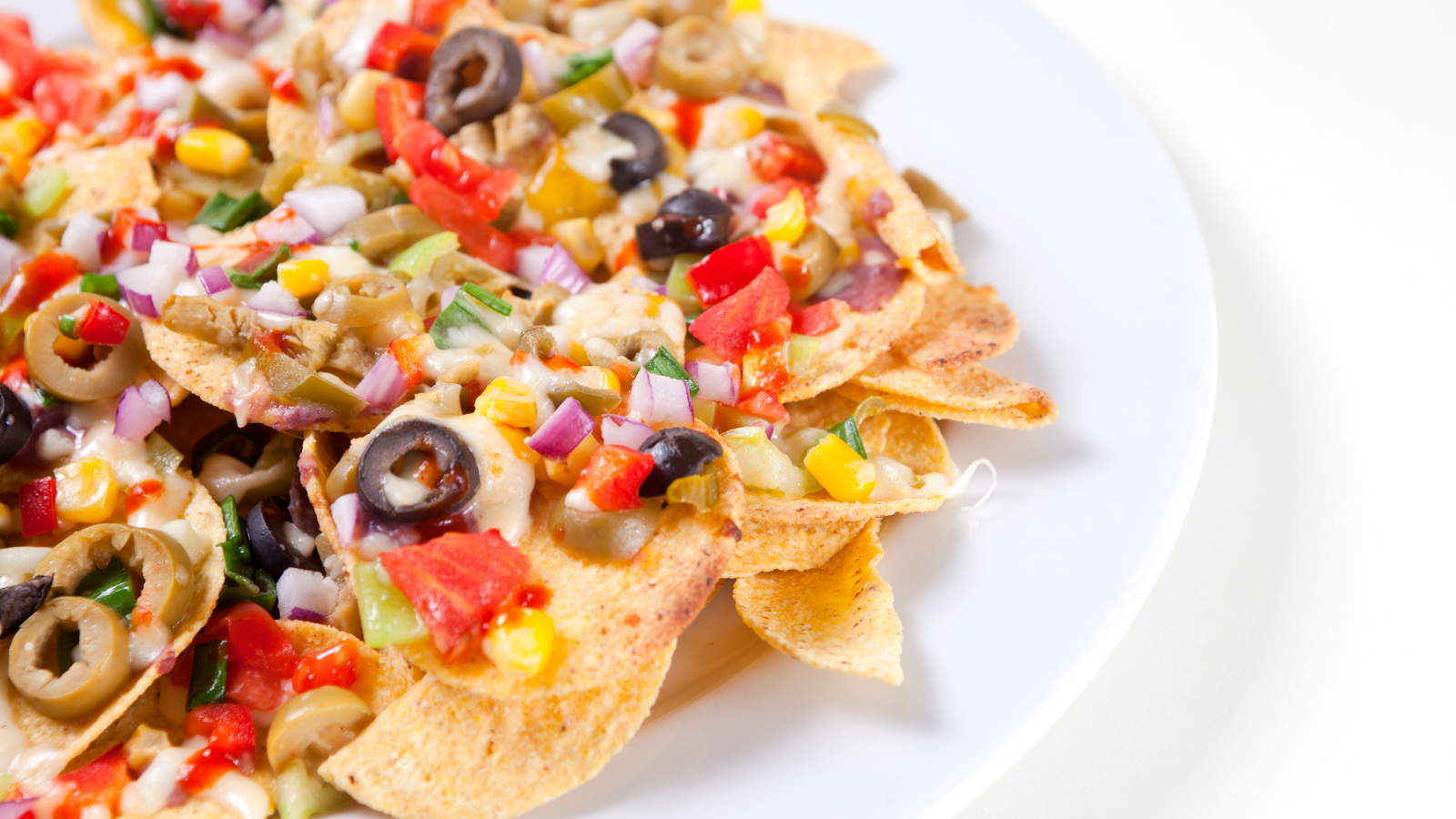 white plate half off the left side of image filled with tortilla chips covered in cheese, olives, corn, tomatoes, and more
