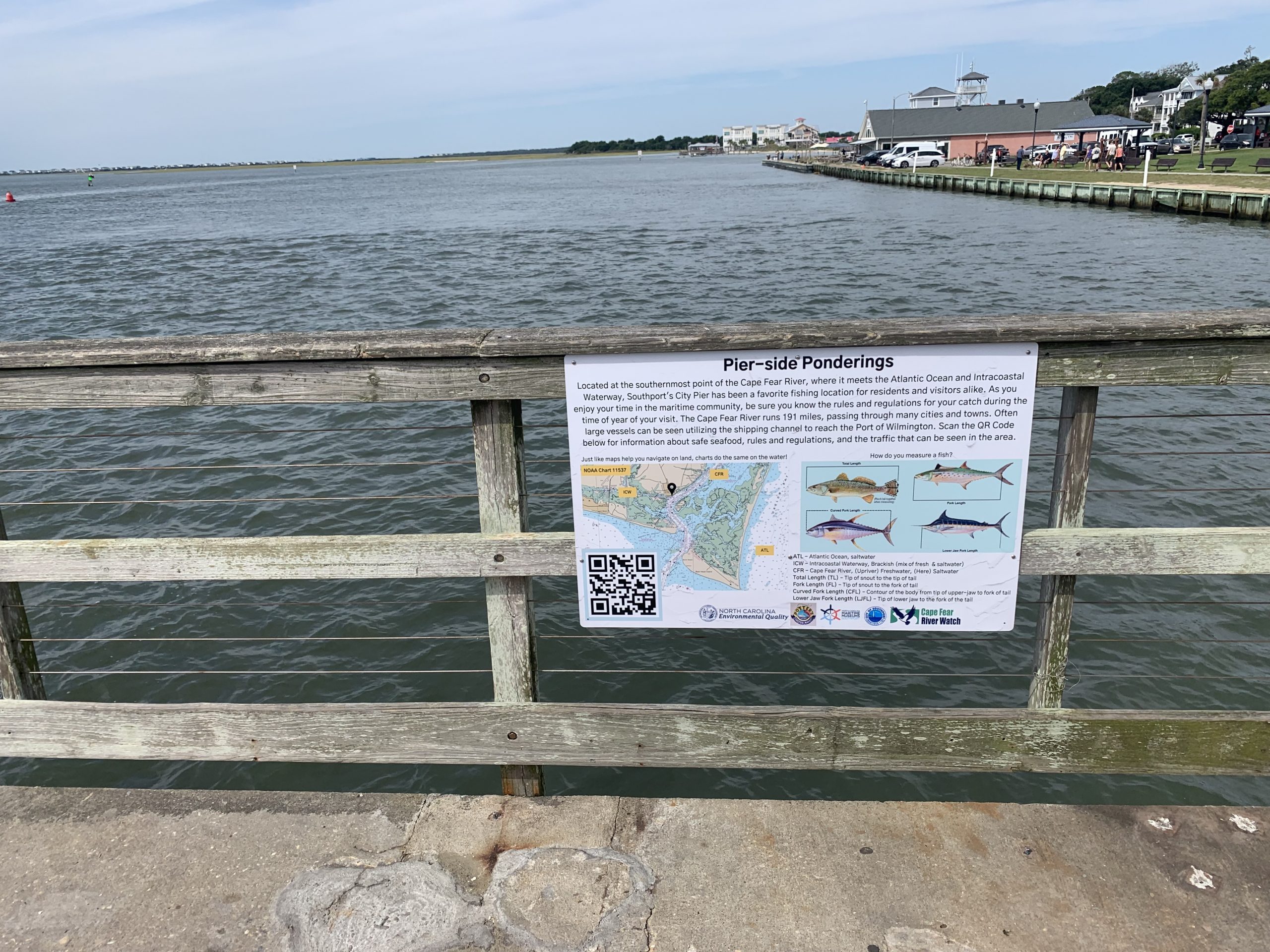 Image Description: White sign containing black font centered at top with image of chart at middle left and image of four fish at middle right. Across bottom is a black and white QR code, logos, and definitions with acronyms. Sign affixed to wooden railings overlooking grey water, bulkheads, and a park in the background