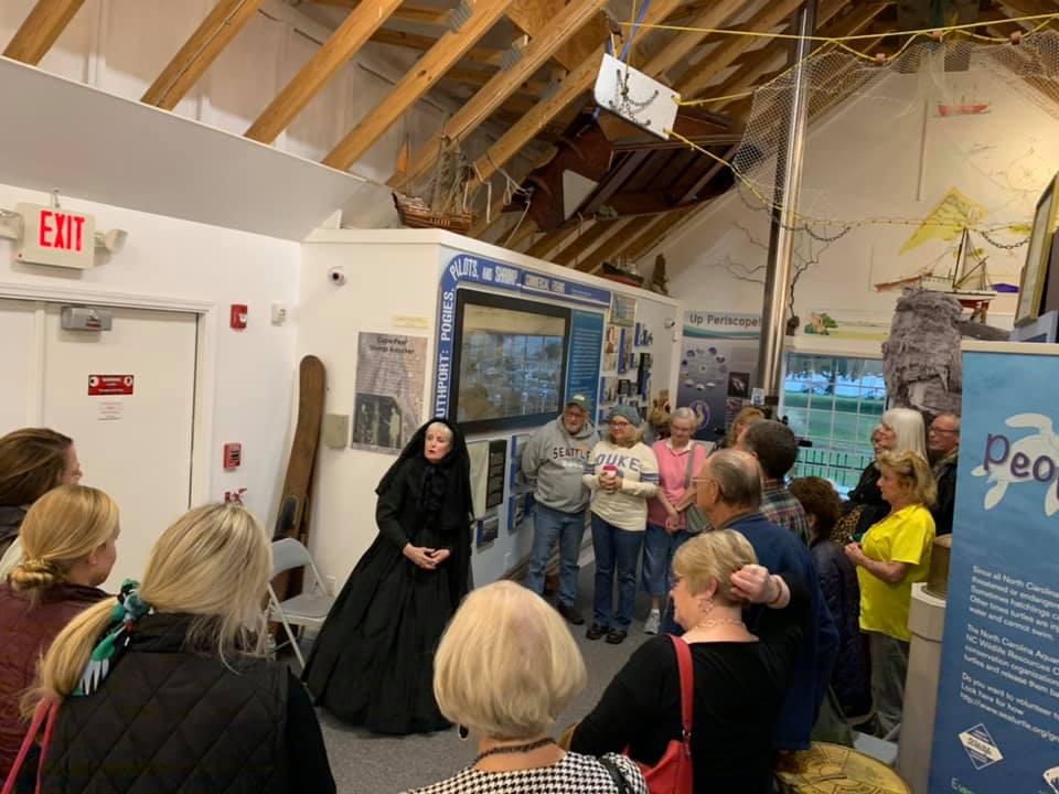 Woman in black historic costume addresses a crowd of people in the museum gallery