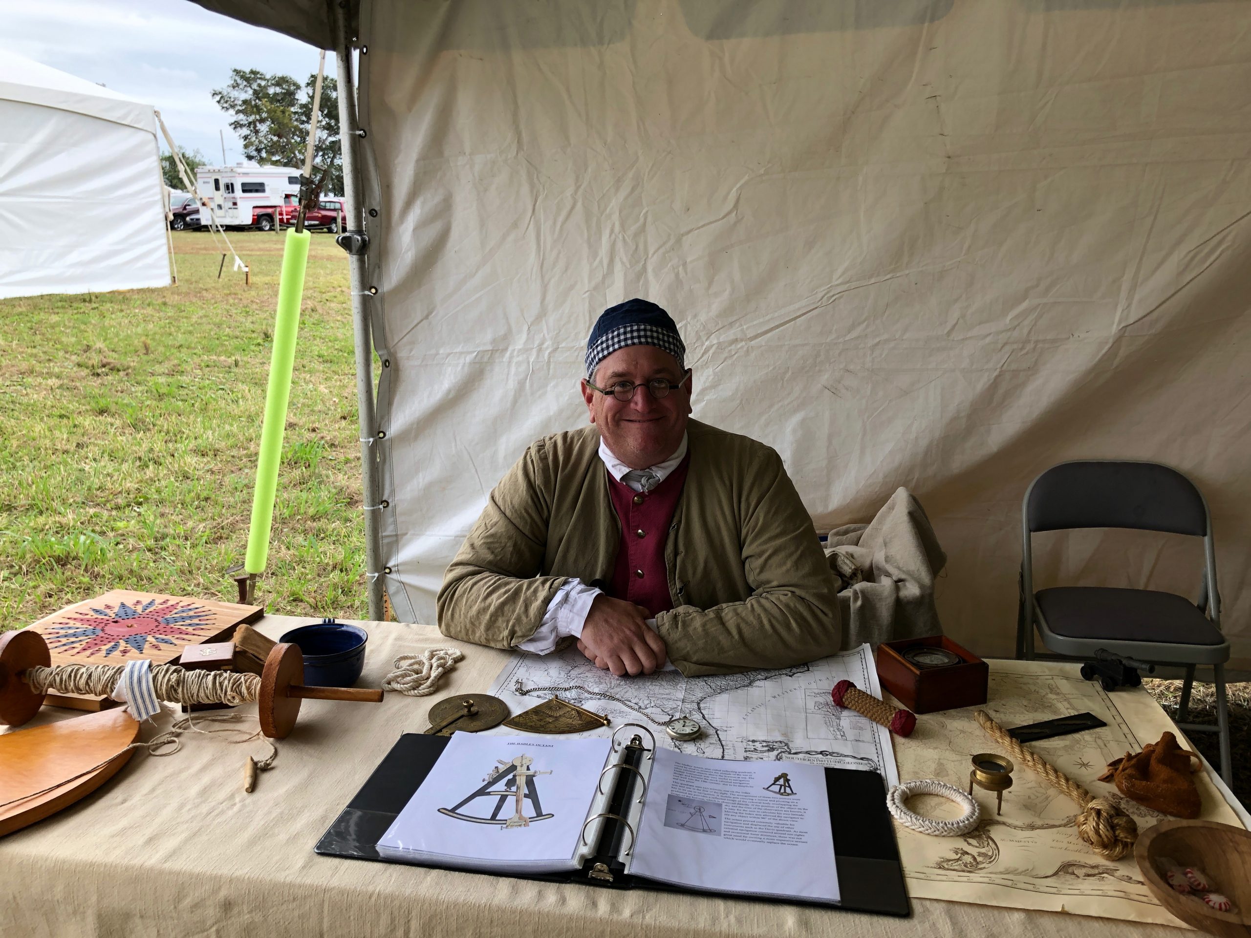 Man in period dress sits smiling and facing camera while sitting at table laden with a variety of historic maritime navigational tools